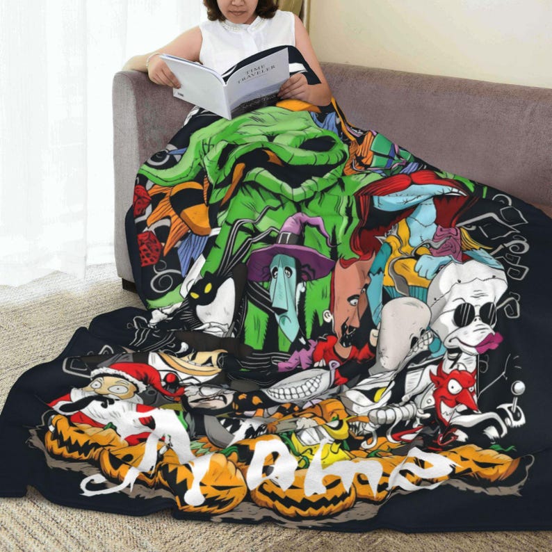 Op de afbeelding: Een zwarte deken met een kleurrijke collage van stripfiguren, waaronder pompoenen, skeletten en een groen monster. De deken is over een bank gedrapeerd. Een persoon leest een boek en gebruikt de deken. De deken heeft een zwarte rand.