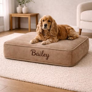May include: A rectangular dog bed in a tan colour, embroidered with the name "Bailey". A golden Cocker Spaniel is resting on the bed. The bed has a contrasting border and is placed on a light-coloured rug.