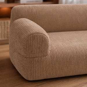 K&ouml;nnte beinhalten: Nahaufnahme eines beigefarbenen Cord-Sofas mit abgerundeter Armlehne. Das Sofa hat eine gerippte Textur und eine neutrale Farbe, passend f&uuml;r verschiedene Einrichtungsstile. Die Armlehne ist geschwungen und gut definiert.