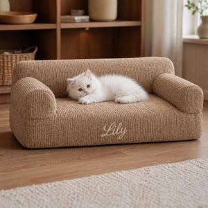K&ouml;nnte beinhalten: Ein kleines Haustiersofa aus beigefarbenem Cordstoff. Das Sofa hat eine abgerundete R&uuml;ckenlehne und Armlehnen. Eine flauschige, wei&szlig;e Katze liegt auf dem Sofa. Der Name "Lily" ist in Schreibschrift auf die Vorderseite des Sofas gestickt.
