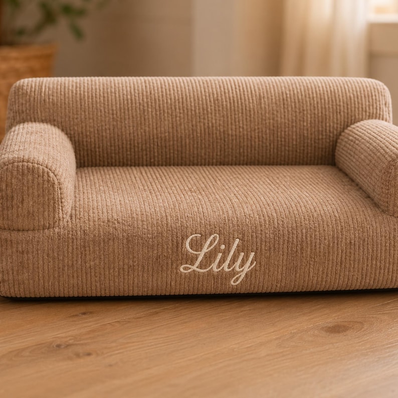 K&ouml;nnte beinhalten: Kleines Hundesofa aus beigefarbenem Cord. Es hat eine abgerundete R&uuml;ckenlehne und Armlehnen, mit dem Namen "Lily" in wei&szlig;er Schrift auf der Vorderseite gestickt. F&uuml;r Haustiere.