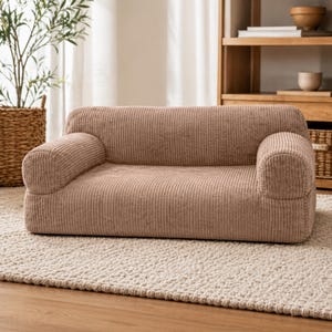 K&ouml;nnte beinhalten: Ein niedriges Sofa aus Cordstoff in einem neutralen Beigeton. Das Sofa hat abgerundete Armlehnen und eine rechteckige Basis, die f&uuml;r einen gem&uuml;tlichen Sitzbereich konzipiert ist. Es steht auf einem hellfarbigen Teppich, was auf eine Wohnzimmereinrichtung hindeutet.