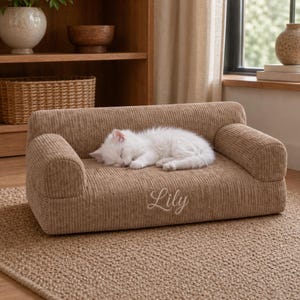 K&ouml;nnte beinhalten: Ein kleines Haustiersofa aus beigefarbenem Cord. Eine flauschige, wei&szlig;e Katze schl&auml;ft auf dem Sofa. Der Name "Lily" ist in Schreibschrift auf die Vorderseite gestickt. Das Sofa steht auf einem gewebten Teppich.