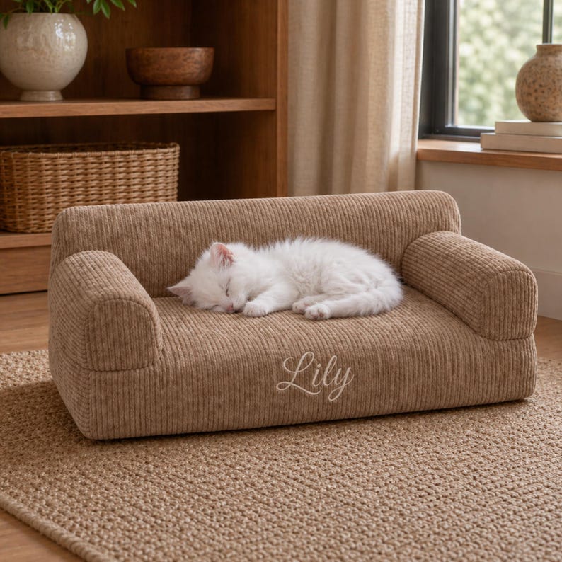 K&ouml;nnte beinhalten: Ein kleines Haustiersofa aus beigefarbenem Cord. Eine flauschige, wei&szlig;e Katze schl&auml;ft auf dem Sofa. Der Name "Lily" ist in Schreibschrift auf die Vorderseite gestickt. Das Sofa steht auf einem gewebten Teppich.
