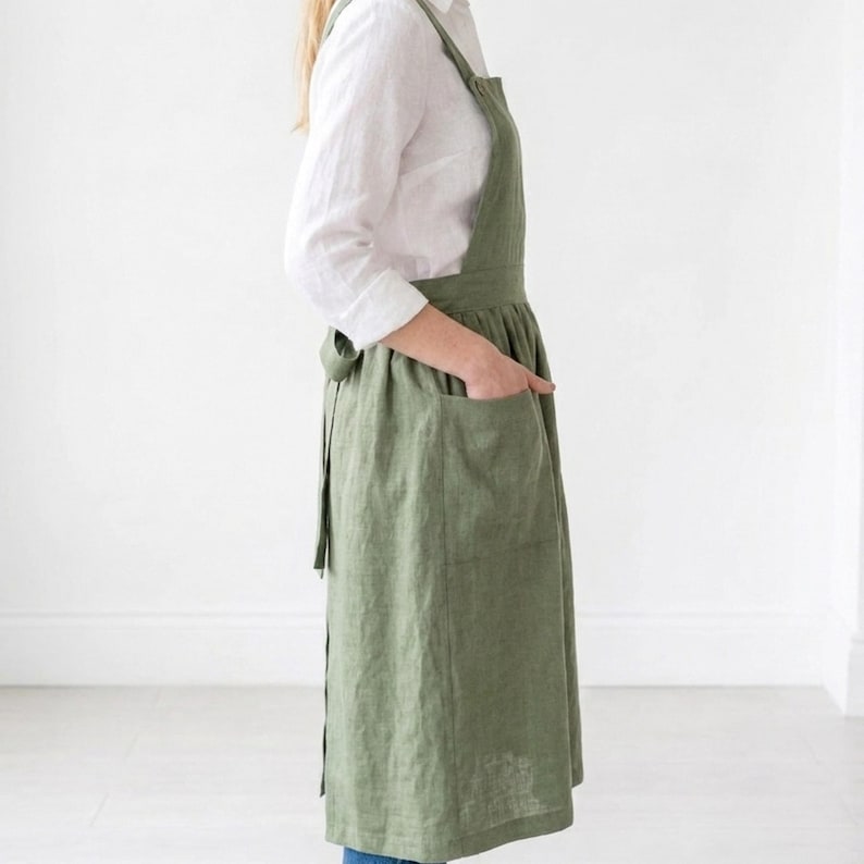 May include: An olive green apron with a cross-back design and two large front pockets. The apron is made of a textured fabric and has a tie closure at the back. The apron is worn over a white long-sleeved shirt.