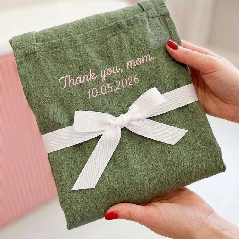 May include: An olive green linen gift bag tied with a white ribbon. The bag has the embroidered text "Thank you, mom. 10.05.2026" in a delicate script. The bag is held by a person with red nail polish.