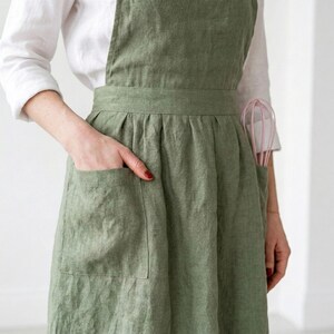 May include: A sage green linen apron with two large front pockets. A pink whisk is tucked into the right pocket. The apron is worn over a white long-sleeved shirt. The apron has a simple, classic design.