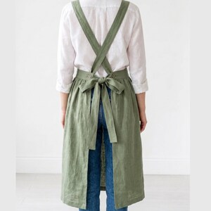May include: A sage green linen apron with cross-back straps and a waist tie. The apron is knee-length and has a full skirt. The model is wearing a white shirt and blue jeans. The apron is designed for kitchen or craft use.