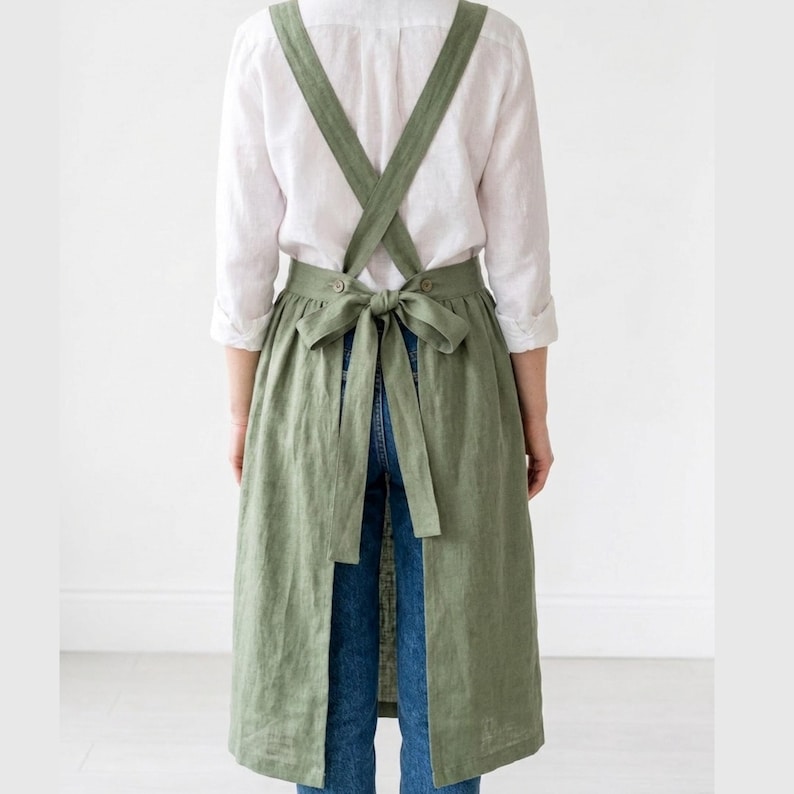 May include: A sage green linen apron with cross-back straps and a waist tie. The apron is knee-length and has a full skirt. The model is wearing a white shirt and blue jeans. The apron is designed for kitchen or craft use.