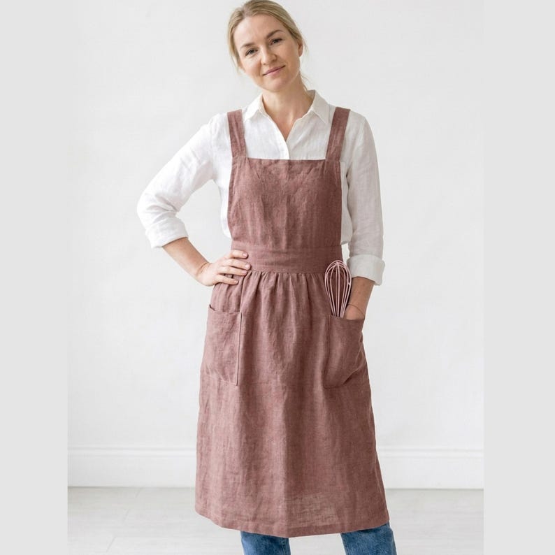 May include: A dusty rose linen apron with a cross-back design, featuring two large front pockets and a waist tie. A pink whisk is tucked into one of the pockets. The apron is worn over a white button-down shirt and blue jeans.