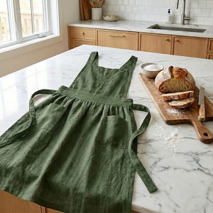 May include: An olive green linen apron with two front pockets and long waist ties. The apron is displayed on a marble countertop next to a wooden cutting board with sliced bread and a knife. A bowl of salt is also present.