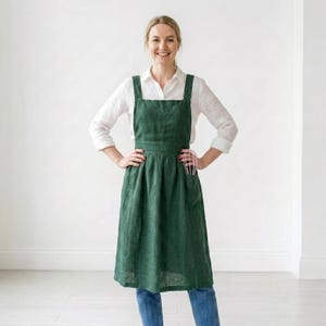 May include: A dark green linen apron with a cross-back design. The apron has a large front pocket and is worn over a white button-down shirt and blue jeans. A pink whisk is in the pocket. The apron is knee-length.