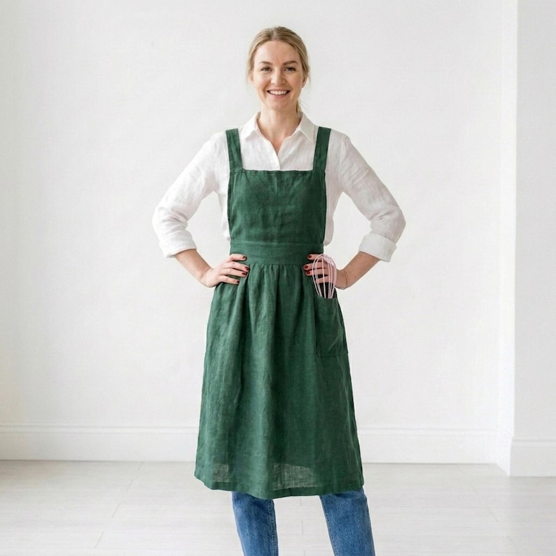 May include: A dark green linen apron with a cross-back design. The apron has a large front pocket and is worn over a white button-down shirt and blue jeans. A pink whisk is in the pocket. The apron is knee-length.