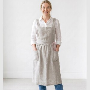 May include: A light gray linen apron with two large front pockets and a pink whisk in the right pocket. The apron is worn over a white shirt and blue jeans. The apron has a cross-back design.