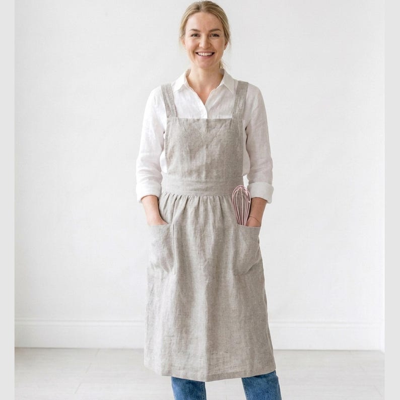 May include: A light gray linen apron with two large front pockets and a pink whisk in the right pocket. The apron is worn over a white shirt and blue jeans. The apron has a cross-back design.