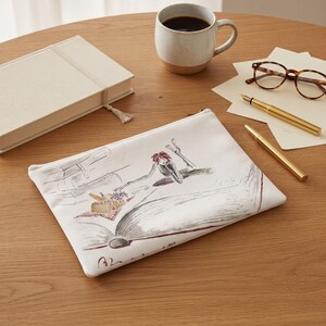 May include: A white rectangular zippered pouch featuring a detailed ink-style illustration of a person reclining on an open book, reaching toward a small picnic basket with grapes. The design includes subtle red and brown accents on the figure and food.