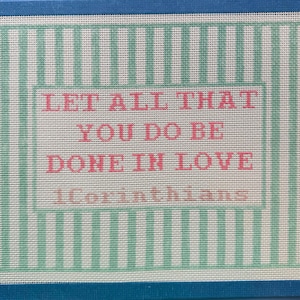 May include: A rectangular needlepoint canvas featuring a light green and white vertical striped border. The center contains the pink text LET ALL THAT YOU DO BE DONE IN LOVE followed by the smaller pink text 1Corinthians on a cream background.