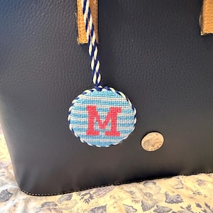 May include: A navy blue leather tote bag with a decorative, round, embroidered charm. The charm features a light blue and white striped background with a red letter "M". The charm is attached to the bag with a blue and white braided cord.