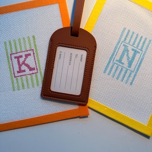 May include: A brown luggage tag with a clear window and printed fields for name, address, telephone, and email, positioned over two square needlepoint canvases with orange and yellow borders featuring the letters K and N in striped patterns.