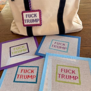 May include: A cream canvas tote bag with black straps and a hanging needlepoint tag reading FUCK TRUMP, accompanied by four square needlepoint canvases featuring the same text in various colored borders and lettering, including purple, blue, and green.