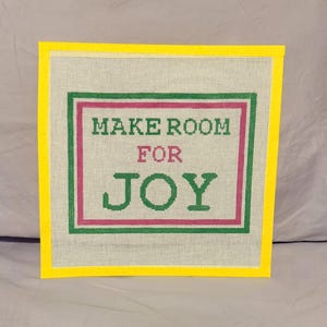 May include: A square needlepoint canvas featuring a bright yellow border and a central design with the text MAKE ROOM FOR JOY. The words are printed in green and pink, surrounded by a green and pink rectangular frame on a white grid background.