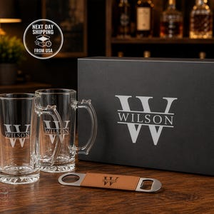 May include: Two clear glass beer mugs and a metal bottle opener with a tan leather grip, all featuring the engraved name WILSON and a large letter W. These items are presented alongside a large black rectangular gift box with the same logo.