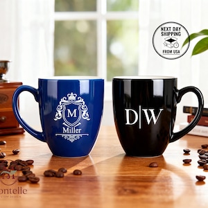May include: Two ceramic mugs, one navy blue and one black, sit on a wooden surface. The blue mug features a white crest with the letter "M" and the name "Miller." The black mug displays the initials "D|W." Coffee beans are scattered around.