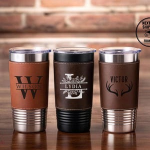 May include: Three stainless steel tumblers with leather-like wraps. The left is tan with a W and WILSON monogram. The center is dark brown with an L, floral design, and LYDIA. The right is dark brown with VICTOR and deer antlers engraved.