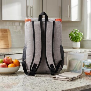 May include: A grey insulated backpack with black straps and orange trim. The backpack has a mesh side pocket and adjustable straps. A bowl of fruit, a folded cloth, and food containers are nearby.