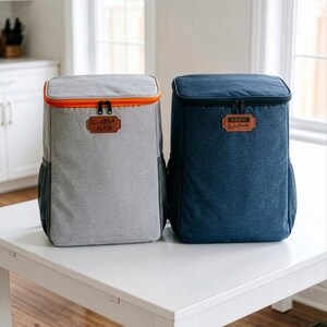 May include: Two insulated cooler backpacks. One is grey with an orange trim and the name "Alice" on a leather patch. The other is navy blue with the name "David Walkins" on a leather patch. Both have black mesh side pockets and black zips.