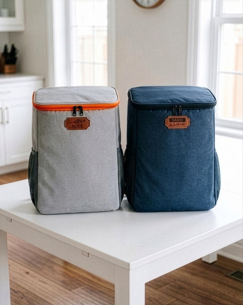May include: Two insulated cooler backpacks. One is grey with an orange trim and the name "Alice" on a leather patch. The other is navy blue with the name "David Walkins" on a leather patch. Both have black mesh side pockets and black zips.