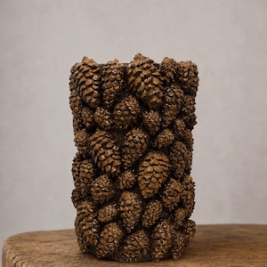 May include: A cylindrical vase covered in a dense arrangement of small, brown, textured pinecones. The exterior surface is entirely composed of these natural elements, creating a uniform, rugged, and organic appearance across the entire structure.