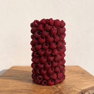 Puede incluir: Una vela cilíndrica con una superficie texturizada cubierta completamente por formas realistas de frambuesas de color rojo intenso. El objeto tiene una estructura vertical uniforme con una disposición densa de bayas individuales, creando una apariencia táctil.