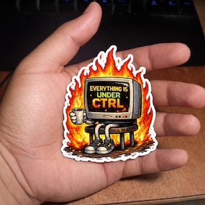 May include: A sticker depicting a retro computer monitor in flames, holding a coffee cup. The monitor shows the text "EVERYTHING IS UNDER CTRL". The sticker has a white border and is held in a hand.