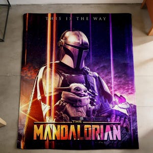May include: A rectangular blanket with a graphic from "The Mandalorian." The image depicts the Mandalorian and Grogu, with the text "THIS IS THE WAY" and "THE MANDALORIAN." The colour scheme includes purple, orange, and black.