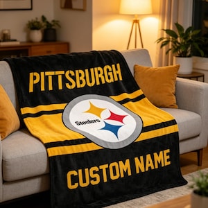 Może przedstawiać: Czarno-złoty koc z napisem "PITTSBURGH" i "CUSTOM NAME" w żółtych literach. Koc posiada logo Pittsburgh Steelers. Koc jest narzucony na szarą sofę.