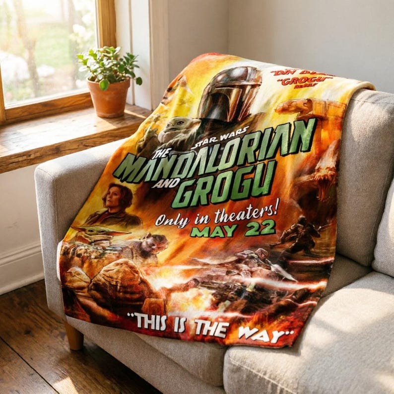 May include: A Star Wars "The Mandalorian and Grogu" throw blanket. The blanket features a movie poster design with the Mandalorian helmet, Grogu, and the text "This is the Way". The blanket is draped over a grey sofa.