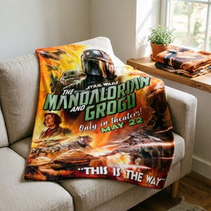 May include: A Star Wars "The Mandalorian and Grogu" throw blanket. The blanket features a movie poster design with the text "THIS IS THE WAY" and "Only in theaters! MAY 22". The blanket is draped over a grey sofa.
