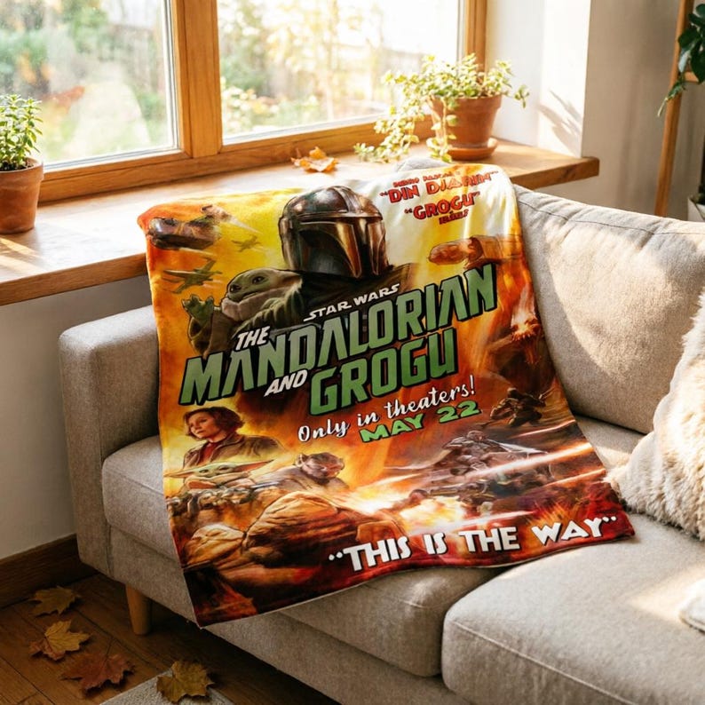 May include: A Star Wars themed blanket with "The Mandalorian and Grogu" movie poster art. The design includes the Mandalorian's helmet, Grogu, and the text "Only in theaters! MAY 22" and "THIS IS THE WAY". The blanket is draped over a sofa.