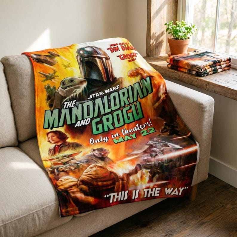 May include: A Star Wars themed blanket featuring The Mandalorian and Grogu. The blanket has a movie poster design with the text "The Mandalorian and Grogu" and "Only in theaters! May 22". The blanket is draped over a light-coloured sofa.