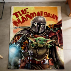 May include: A rectangular throw blanket featuring an illustration of The Mandalorian and Grogu from Star Wars. The blanket has a tan background with the words "THE MANDALORIAN" in red and yellow. The Mandalorian is in his armour, holding a weapon, with Grogu in a carrier.