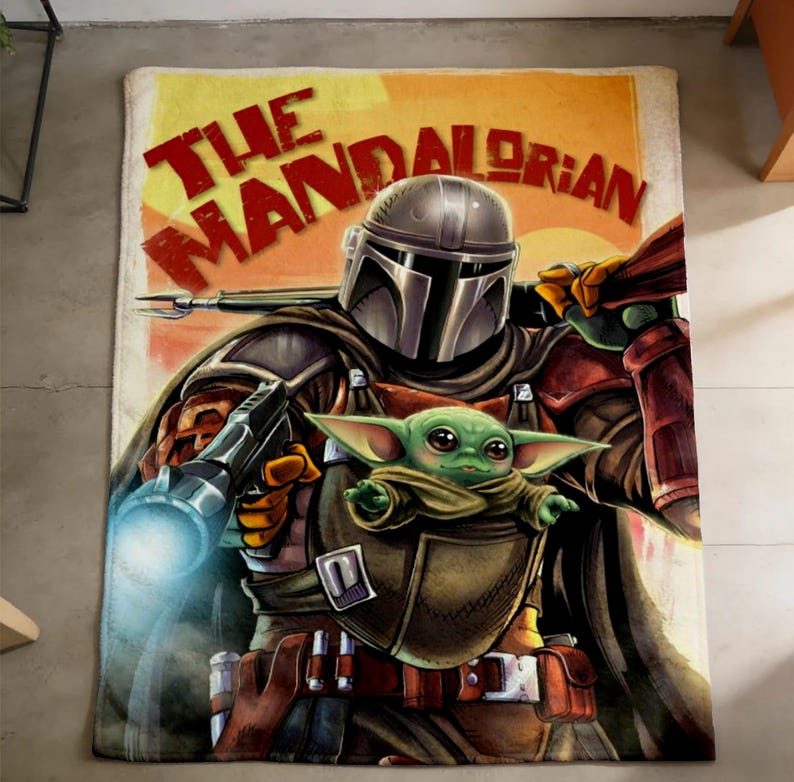 May include: A rectangular throw blanket featuring an illustration of The Mandalorian and Grogu from Star Wars. The blanket has a tan background with the words "THE MANDALORIAN" in red and yellow. The Mandalorian is in his armour, holding a weapon, with Grogu in a carrier.