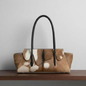 Könnte beinhalten: Eine strukturierte Handtasche mit braun-weißem Kuhfellmuster und strukturierter Felloberfläche. Die Tasche hat eine horizontale, rechteckige Form mit Klappverschluss und zwei hohen, dünnen Tragegriffen aus schwarzem Leder.