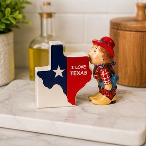 May include: A ceramic salt and pepper shaker set featuring a Texas-shaped piece with the blue, white, and red state flag design and the text I LOVE TEXAS, paired with a figurine of a person in a red hat, plaid shirt, and cowboy boots.
