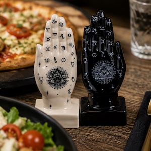 May include: Two ceramic hand sculptures standing on square bases, one in white and one in black. Each hand features intricate black or white line art, including zodiac symbols, celestial icons, and a central eye of providence design.