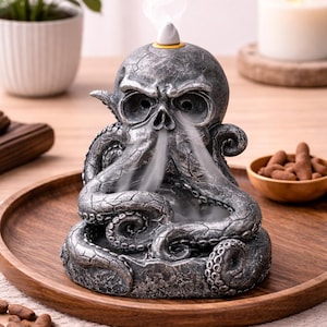 May include: A detailed, gray resin incense burner shaped like a skull with octopus tentacles. Smoke rises from the skull's eye sockets and mouth. The burner sits on a wooden tray, with a small candle and plant in the background.