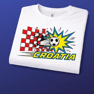 Puede incluir: Una camiseta blanca con un gr&aacute;fico de un bal&oacute;n de f&uacute;tbol atravesando un muro a cuadros rojos y blancos. El dise&ntilde;o incluye un efecto de explosi&oacute;n amarillo y azul detr&aacute;s del bal&oacute;n y la palabra CROATIA en letras amarillas con borde azul.