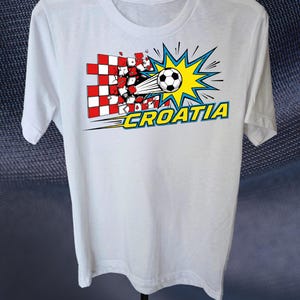 Puede incluir: Una camiseta blanca de manga corta con un gr&aacute;fico de un bal&oacute;n de f&uacute;tbol atravesando una pared de cuadros rojos y blancos. El dise&ntilde;o incluye un fondo de estrella amarilla y la palabra CROATIA en letras amarillas con un borde azul.