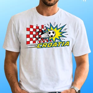 Puede incluir: Una camiseta blanca con un gr&aacute;fico de un bal&oacute;n de f&uacute;tbol atravesando una pared de cuadros rojos y blancos. El dise&ntilde;o incluye un efecto de explosi&oacute;n amarilla y la palabra CROATIA en letras amarillas negritas con borde azul al frente.