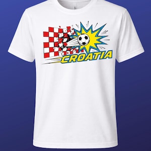 Puede incluir: Una camiseta blanca de manga corta con un gr&aacute;fico de un bal&oacute;n de f&uacute;tbol atravesando una pared a cuadros rojos y blancos. Detr&aacute;s del bal&oacute;n hay un dise&ntilde;o de estrella amarilla y azul. La palabra CROATIA est&aacute; impresa en amarillo con borde azul.